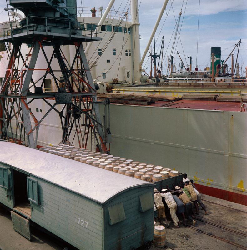 Color photograph of a group of men pushing a train car filled with barrels next to a ship