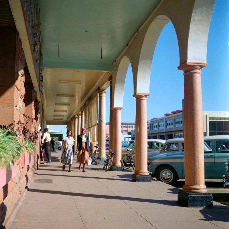 Color image of two woman walking down a outdoor promenade with arches along the right side