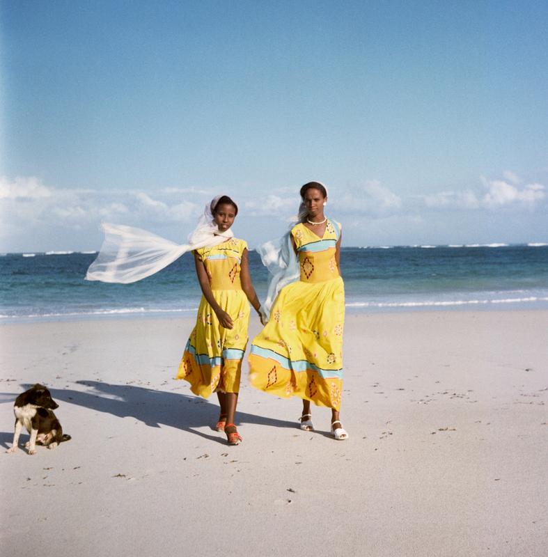 Color image of two girls wearing yellow dresses on a beach; a black and white dog in the LL corner